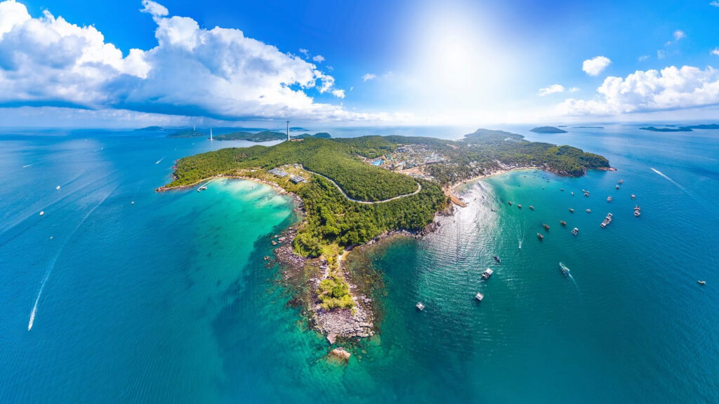 Aerial view of lush green Hon Thom Phu Quoc island surrounded by turquoise ocean, with boats scattered in the water and a winding road under a bright, sunny sky.