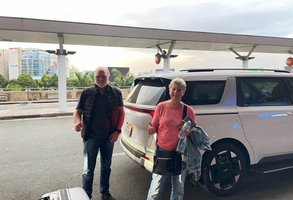Two people with luggage stand beside a white car at an airport pick up service Phu Quoc drop-off area, giving thumbs up. City buildings and greenery are visible in the background under a cloudy sky.